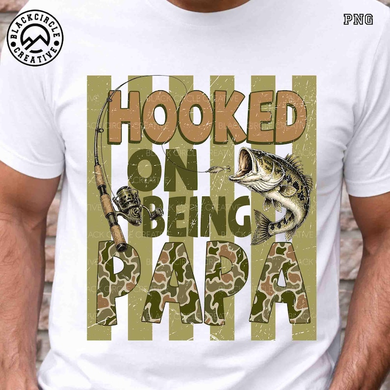 Op de afbeelding: Wit T-shirt met een visserijthema. Het ontwerp toont de tekst "HOOKED ON BEING PAPA" in bruine en camouflage letters, een hengel en een vis met open mond.