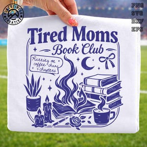 Può includere: T-shirt bianco con un design grafico blu navy. Il design presenta il testo "Tired Moms Book Club" con illustrazioni di libri, candele, una tazza di caffè e una pianta. È inclusa anche la frase "Running on coffee, chaos, + chapters".