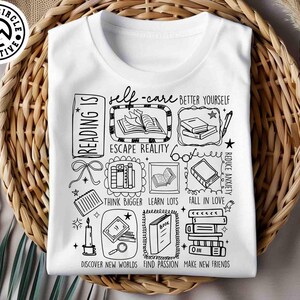 May include: White t-shirt with a black graphic design featuring the words "Reading Is" and various book-related illustrations. The design includes phrases like "Escape Reality" and "Discover New Worlds."