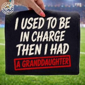 May include: Black t-shirt with white text that reads "I USED TO BE IN CHARGE THEN I HAD A GRANDDAUGHTER" in a distressed font. The text "A GRANDDAUGHTER" is in a red rectangle. The shirt is held up by a hand.