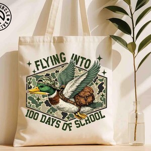 Pu&ograve; includere: Borsa tote color crema con un'anatra dei cartoni animati con cappello e zaino mimetico, con una matita nel becco. Il testo recita "Flying Into 100 Days of School". La borsa ha un logo Black Circle Creative.