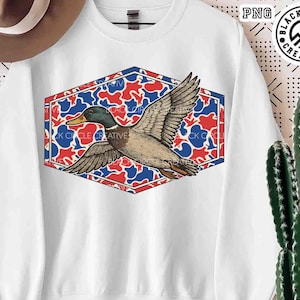 May include: White sweatshirt featuring a detailed illustration of a flying duck. The duck is set against a red, white, and blue camouflage pattern within a hexagonal frame. The sweatshirt has the text "BLACK CIRCLE CREATIVE".