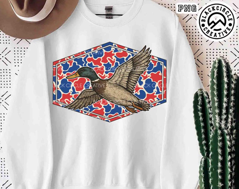 May include: White sweatshirt featuring a detailed illustration of a flying duck. The duck is set against a red, white, and blue camouflage pattern within a hexagonal frame. The sweatshirt has the text "BLACK CIRCLE CREATIVE".