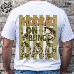 Op de afbeelding: Wit T-shirt met een visserij-thema grafisch ontwerp. Het ontwerp toont een hengel, een vis en de tekst "HOOKED ON BEING DAD" in bruine en camouflage letters. Het shirt heeft een ronde hals.