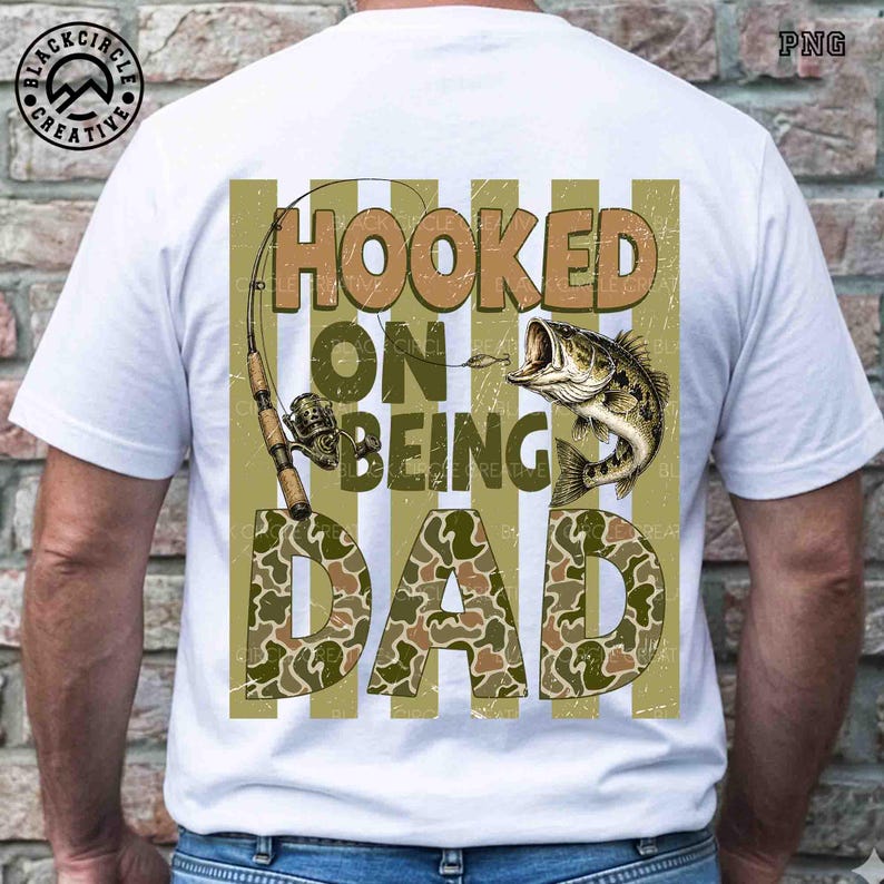 Op de afbeelding: Wit T-shirt met een visserij-thema grafisch ontwerp. Het ontwerp toont een hengel, een vis en de tekst "HOOKED ON BEING DAD" in bruine en camouflage letters. Het shirt heeft een ronde hals.