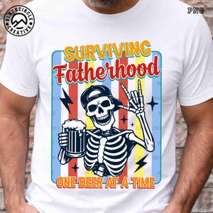 May include: White t-shirt with a graphic of a skeleton wearing a cap, holding a beer mug, and making a rock-and-roll hand gesture. The text reads "SURVIVING Fatherhood ONE BEER AT A TIME."