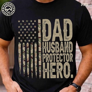Puede incluir: Camiseta negra con un dise&ntilde;o de bandera americana de camuflaje desgastado. La camiseta presenta las palabras "DAD HUSBAND PROTECTOR HERO" en una fuente beige texturizada. Se ve un reloj en la mu&ntilde;eca.