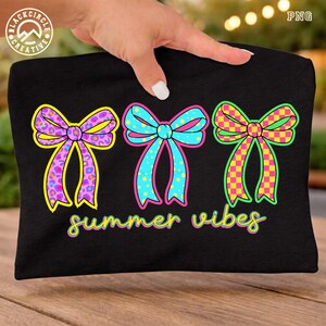 May include: Black t-shirt featuring three colorful bow designs. The bows have unique patterns, including leopard print, polka dots, and a checkered design. The text "summer vibes" is written in a playful font below the bows.