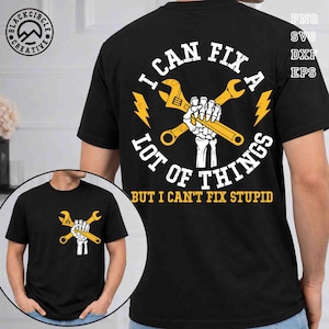 Puede incluir: Camiseta negra con un gráfico que presenta una mano esquelética sosteniendo llaves cruzadas, con el texto "I CAN FIX A LOT OF THINGS BUT I CAN'T FIX STUPID". El diseño incluye rayos amarillos.