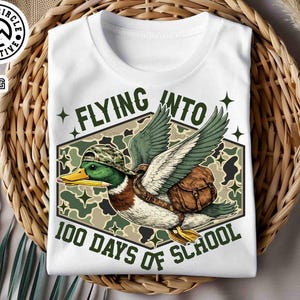 Pu&ograve; includere: T-shirt bianca con un'anatra dei cartoni animati con cappello e zaino mimetico, che tiene una matita. La maglietta ha la scritta "FLYING INTO 100 DAYS OF SCHOOL" in verde. La maglietta &egrave; su un cestino intrecciato.