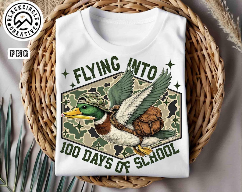 Pu&ograve; includere: T-shirt bianca con un'anatra dei cartoni animati con cappello e zaino mimetico, che tiene una matita. La maglietta ha la scritta "FLYING INTO 100 DAYS OF SCHOOL" in verde. La maglietta &egrave; su un cestino intrecciato.