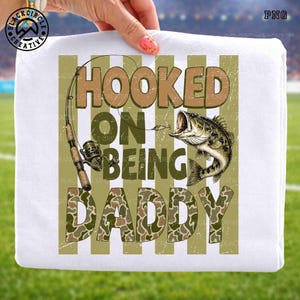 Op de afbeelding: Wit T-shirt met de tekst "HOOKED ON BEING DADDY" in bruine en camouflage letters. Het ontwerp bevat een hengel en een largemouth bass. Het shirt heeft een distressed, vintage uitstraling.