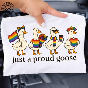 Puede incluir: Camiseta blanca con un diseño de cuatro gansos de dibujos animados, cada uno adornado con accesorios con temática de arcoíris. Un ganso sostiene una bandera arcoíris, mientras que otros llevan bufandas, flores y gafas de sol. El texto "just a proud goose" está impreso debajo.