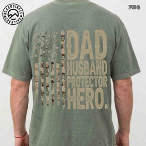 Puede incluir: Camiseta verde salvia con un dise&ntilde;o de bandera estadounidense desgastada con rayas y estrellas de camuflaje. La camiseta muestra las palabras "DAD, HUSBAND, PROTECTOR, HERO" en una fuente beige texturizada. El logotipo de Black Circle Creative es visible.
