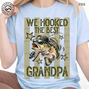 Pode incluir: Camiseta azul clara com um gr&aacute;fico de um peixe usando &oacute;culos de sol e um chap&eacute;u, segurando uma vara de pescar. O texto diz "WE HOOKED THE BEST GRANDPA" em uma fonte desgastada. Estrelas e listras verticais completam o design.