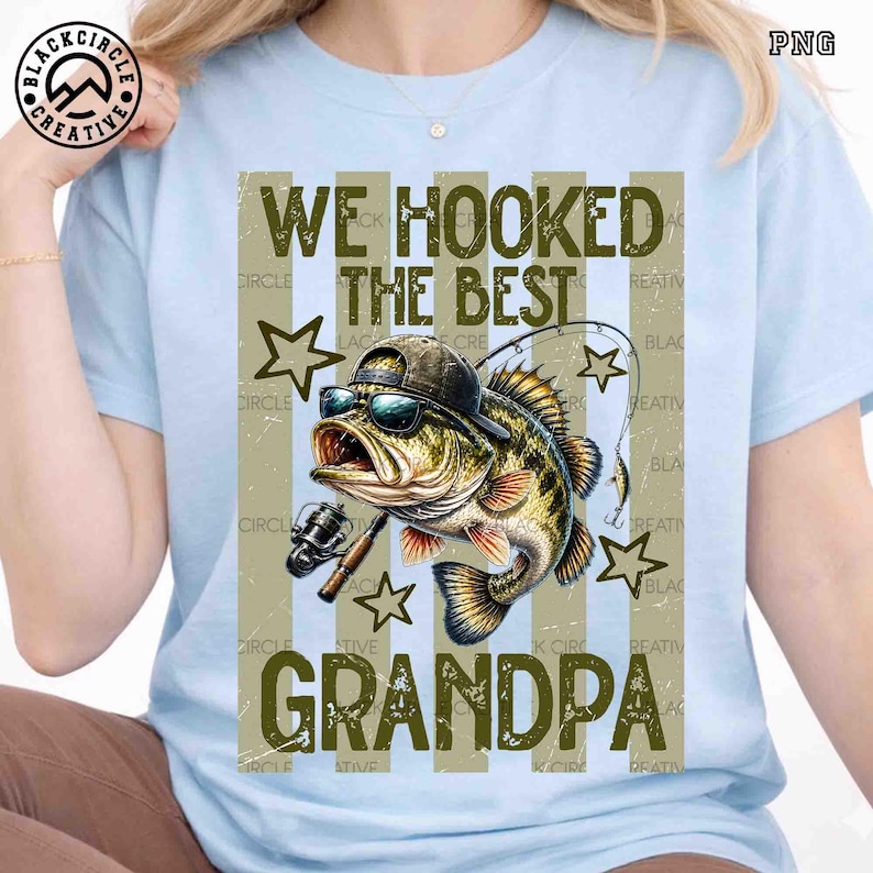 Pode incluir: Camiseta azul clara com um gr&aacute;fico de um peixe usando &oacute;culos de sol e um chap&eacute;u, segurando uma vara de pescar. O texto diz "WE HOOKED THE BEST GRANDPA" em uma fonte desgastada. Estrelas e listras verticais completam o design.