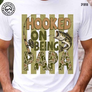 Op de afbeelding: Wit T-shirt met een visserijthema. Het ontwerp toont de woorden "Hooked on Being Papa" in bruin en camouflage, een hengel en een vis. Het T-shirt heeft een distressed look.