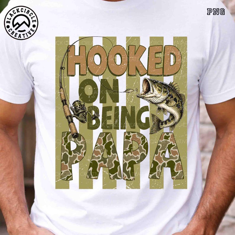 Op de afbeelding: Wit T-shirt met een visserijthema. Het ontwerp toont de woorden "Hooked on Being Papa" in bruin en camouflage, een hengel en een vis. Het T-shirt heeft een distressed look.