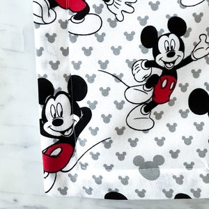May include: White fabric with a repeating pattern of Mickey Mouse in black and red. The fabric has a gray border.