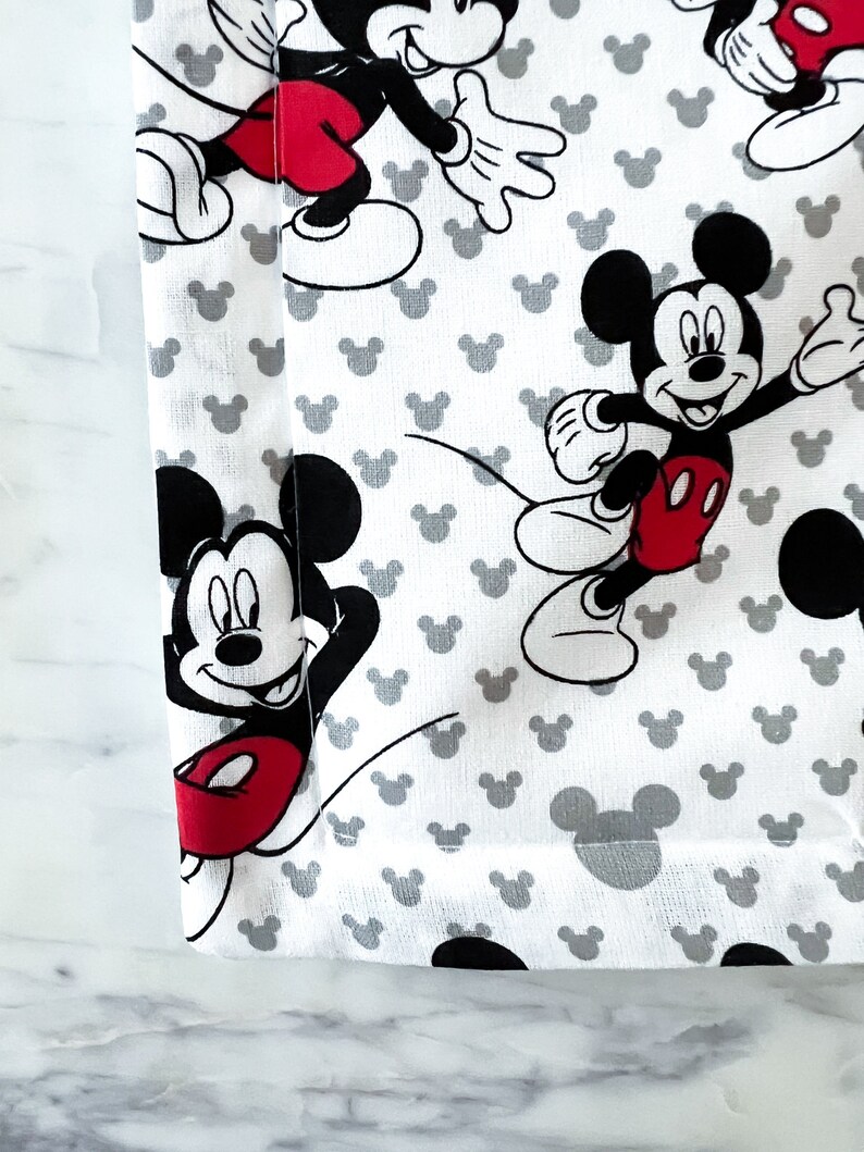 May include: White fabric with a repeating pattern of Mickey Mouse in black and red. The fabric has a gray border.