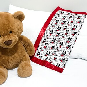 May include: A white and red baby blanket with a Mickey Mouse print. The blanket has a red satin border and is lying on a white bed with a brown teddy bear.