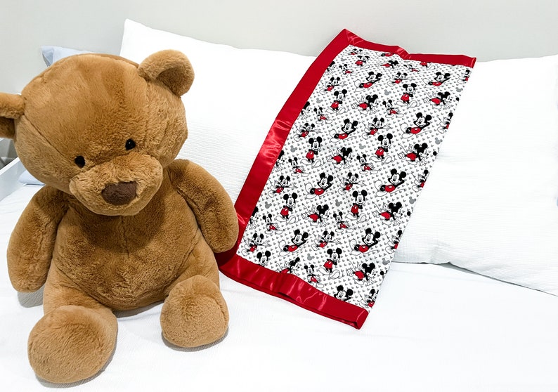 May include: A white and red baby blanket with a Mickey Mouse print. The blanket has a red satin border and is lying on a white bed with a brown teddy bear.