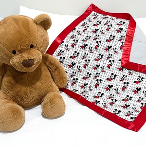 May include: A white and red baby blanket with a Mickey Mouse print. The blanket has a red satin border. A brown teddy bear is sitting on a white bed.