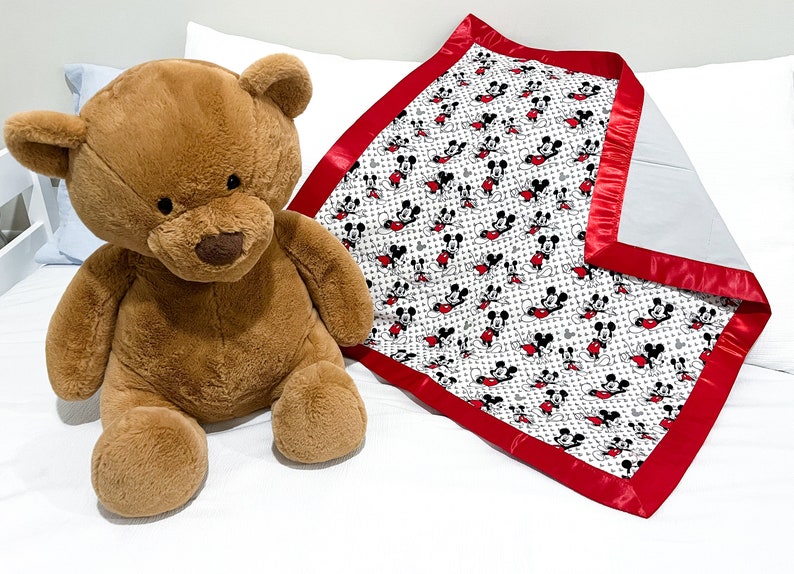 May include: A white and red baby blanket with a Mickey Mouse print. The blanket has a red satin border. A brown teddy bear is sitting on a white bed.
