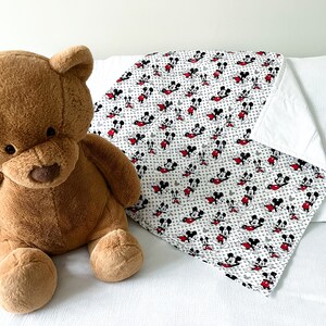 May include: A white and gray Mickey Mouse print blanket with red accents. The blanket is folded and lying on a white surface. A brown teddy bear is sitting in the foreground.