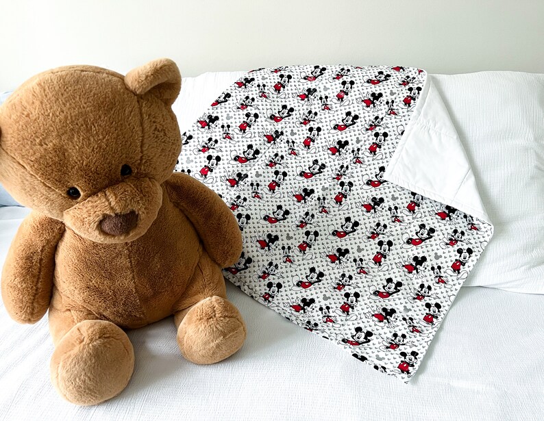 May include: A white and gray Mickey Mouse print blanket with red accents. The blanket is folded and lying on a white surface. A brown teddy bear is sitting in the foreground.