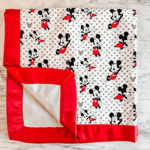 May include: A red and white baby blanket with a Mickey Mouse print. The blanket has a red satin border.