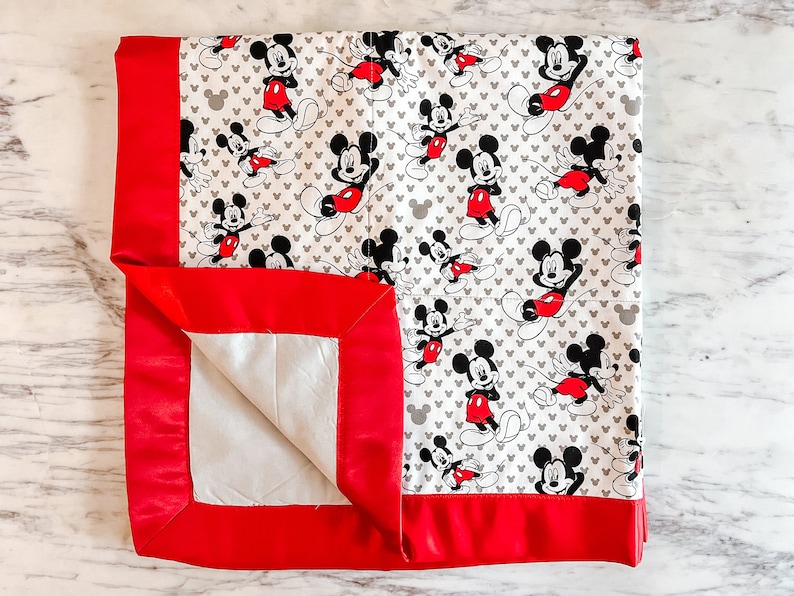 May include: A red and white baby blanket with a Mickey Mouse print. The blanket has a red satin border.