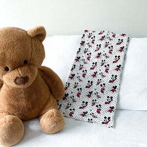 May include: A brown teddy bear sitting on a white bed with a Mickey Mouse print blanket. The blanket is white with red and black Mickey Mouse characters.