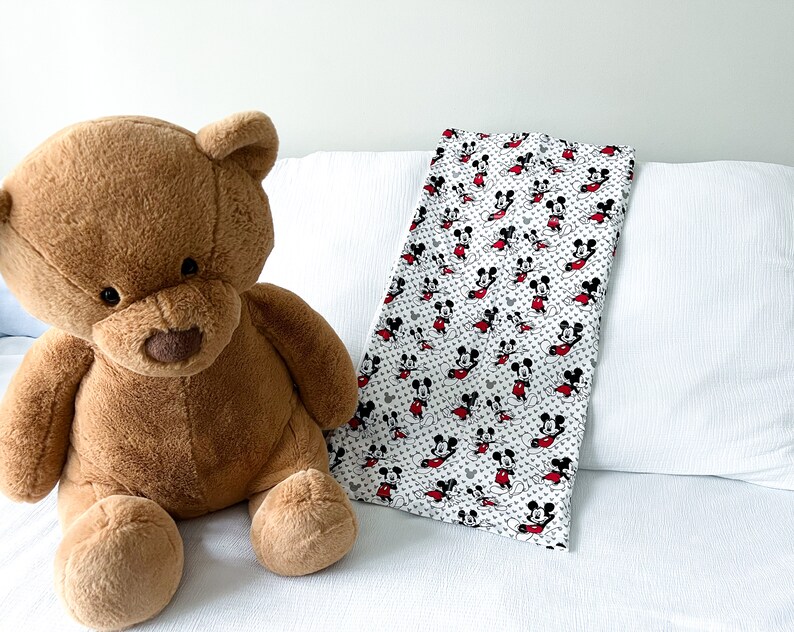 May include: A brown teddy bear sitting on a white bed with a Mickey Mouse print blanket. The blanket is white with red and black Mickey Mouse characters.