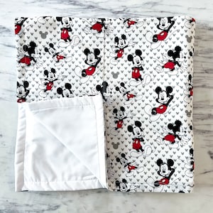 May include: A white and gray baby blanket with a Mickey Mouse print. The blanket is folded in half and the white side is facing up.