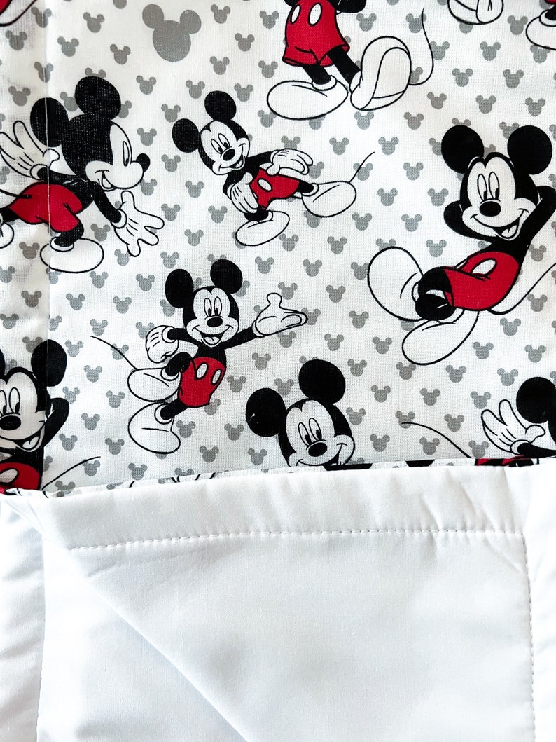 May include: White and gray fabric with a repeating pattern of Mickey Mouse. The fabric has a white backing.