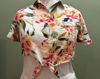 May include: A white cropped button-down shirt with a floral print in shades of pink, yellow, green, and blue. The shirt has short sleeves and a tie at the front.