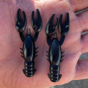 May include: Two black and blue crawfish-shaped fishing lures with a speckled finish.