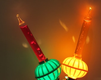 May include: Two vintage Christmas tree light bulbs with green and orange plastic covers. The bulbs are lit and glowing. The bulb on the left has a red liquid inside and the bulb on the right has a clear liquid inside.