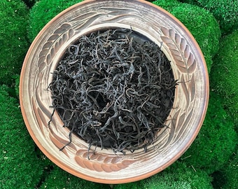 May include: A round ceramic plate with a brown and tan glaze featuring an embossed wheat pattern along the rim. The plate is filled with a mound of dark, dried, twisted tea leaves. The surface of the plate shows a rustic, hand-crafted texture.