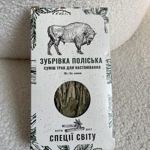 Pode incluir: Caixa retangular de ervas Zubrivka Poliska para infusão. A caixa apresenta uma ilustração a preto e branco de um bisonte e texto em ucraniano. Uma janela circular revela as ervas secas no interior. O texto "ESTD 2017" também é visível.