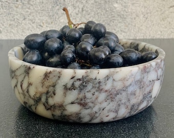May include: A white marble bowl with gray and brown marbling, filled with a bunch of dark purple grapes.