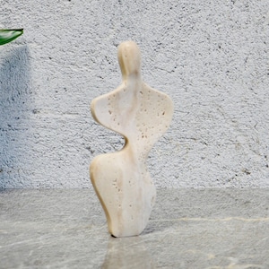 May include: A white abstract sculpture of a person, carved from stone, standing on a gray marble surface.