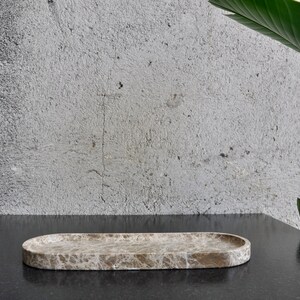May include: A light brown oval marble tray with a textured surface. The tray is sitting on a black surface.