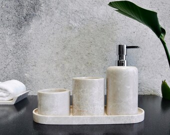 Op de afbeelding: Een set van drie witte marmeren badkameraccessoires, waaronder een zeepdispenser, een beker en een tandenborstelhouder, allemaal op een bijpassende ovale dienblad.