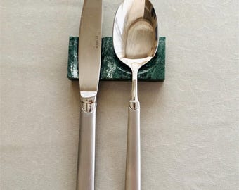 May include: A stainless steel knife and spoon set, resting on a green marble holder. The knife has a curved blade and a long, rectangular handle. The spoon has a rounded bowl and a matching handle. The word "Pols Potten" is visible on the knife.