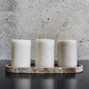May include: Three white and grey pillar candles on a brown marble tray. The candles are arranged in a row, with the white candles at either end and the grey candle in the middle.