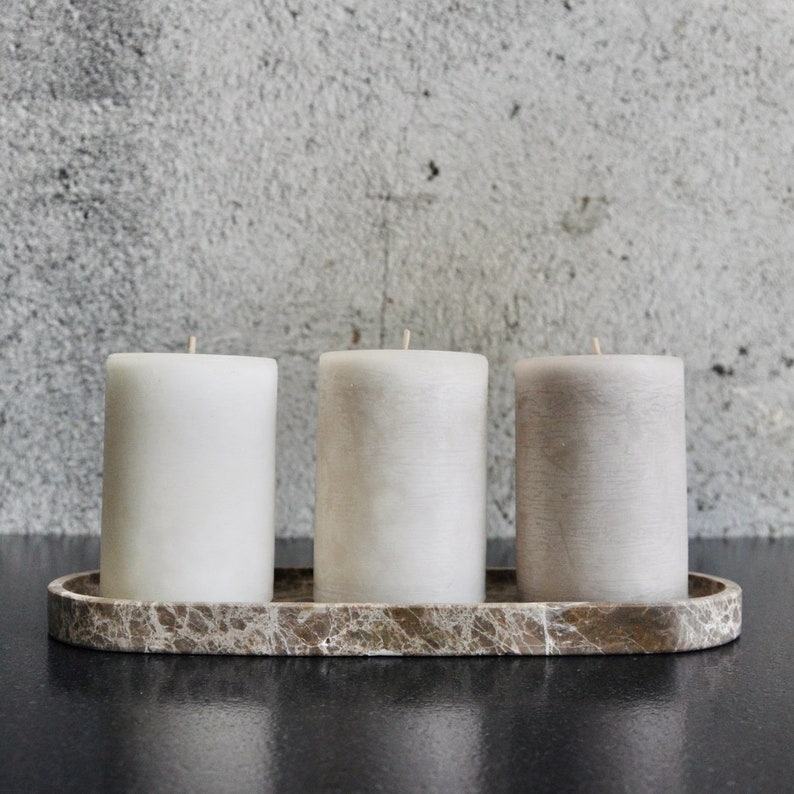 May include: Three white and grey pillar candles on a brown marble tray. The candles are arranged in a row, with the white candles at either end and the grey candle in the middle.