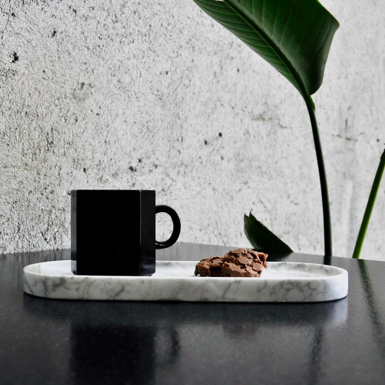 May include: A black ceramic mug and a chocolate chip cookie on a white marble oval serving tray.