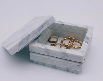 Op de afbeelding: Een wit marmeren juwelendoosje met een open deksel, waarin gouden ringen en oorbellen te zien zijn.